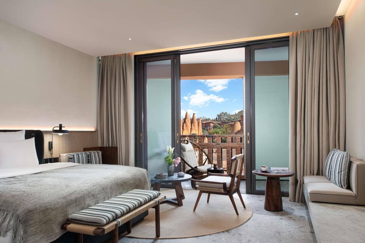A modern hotel room with a large bed, a sofa, a small table, and a balcony with a view of buildings.