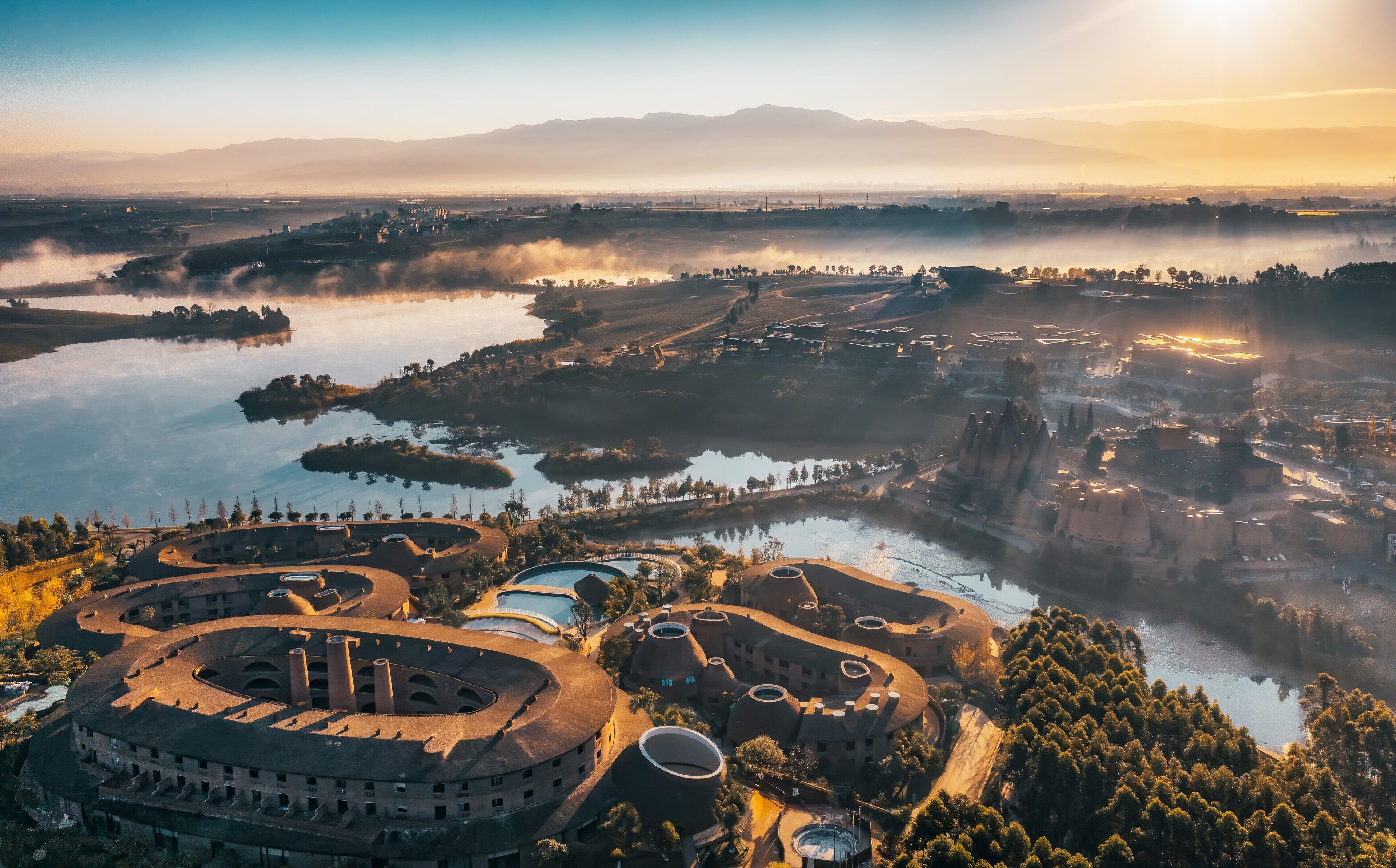 Vista aérea de um resort com edifícios circulares, um lago e montanhas ao redor.