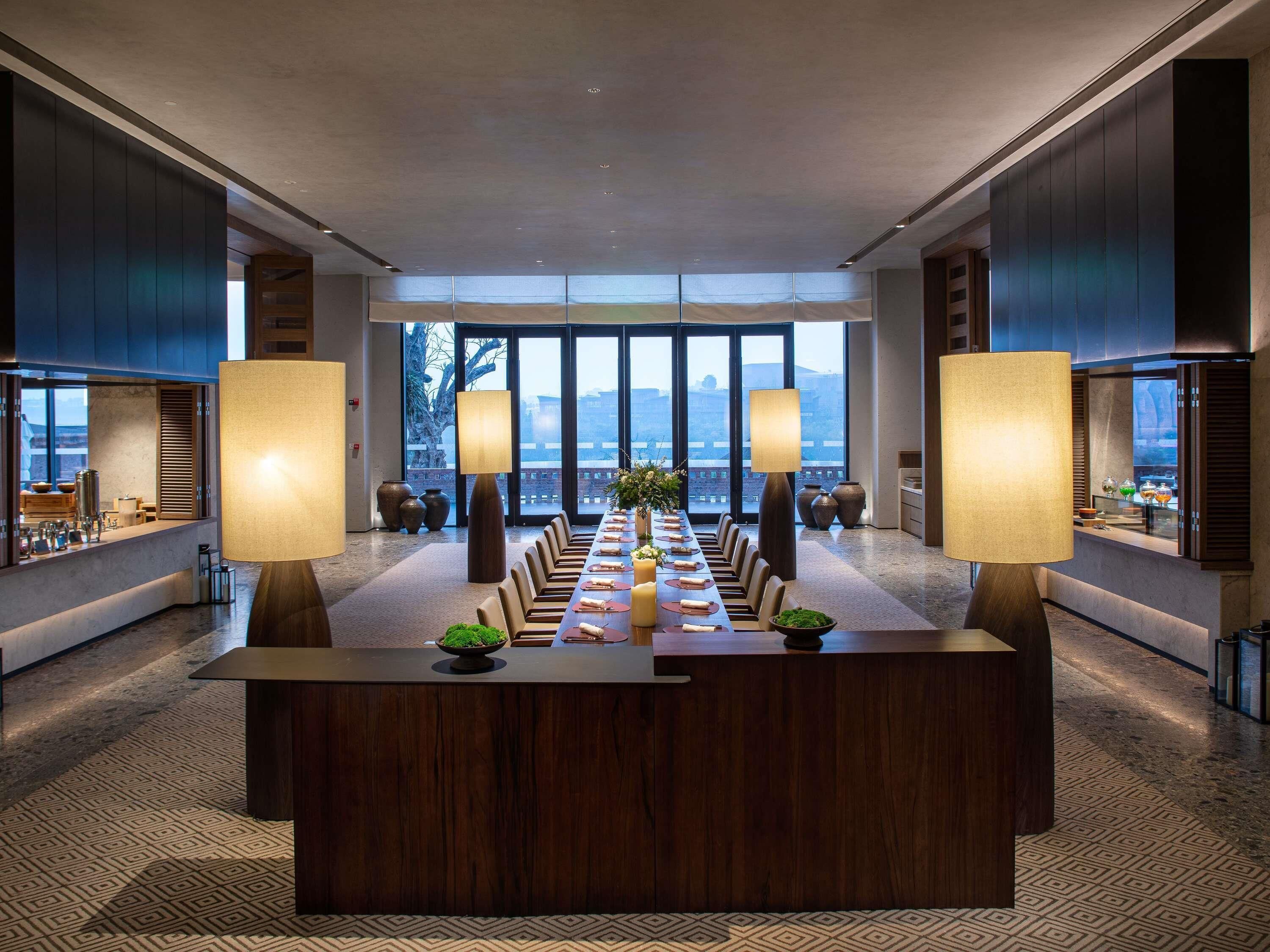 A modern hotel dining area with a long table, two large lamps, and a view of the mountains through large windows.
