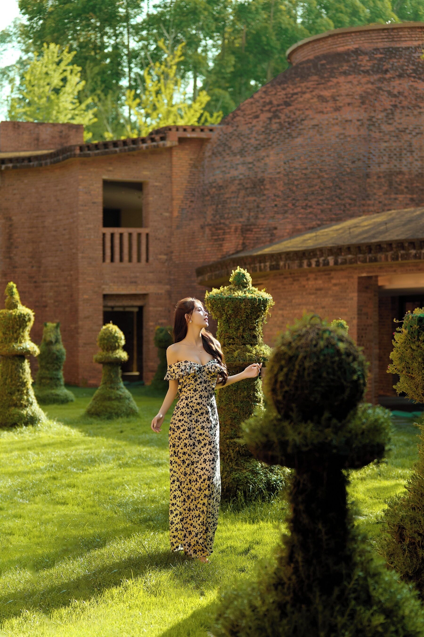 A woman in a patterned dress standing in a well-manicured garden with a brick building in the background.