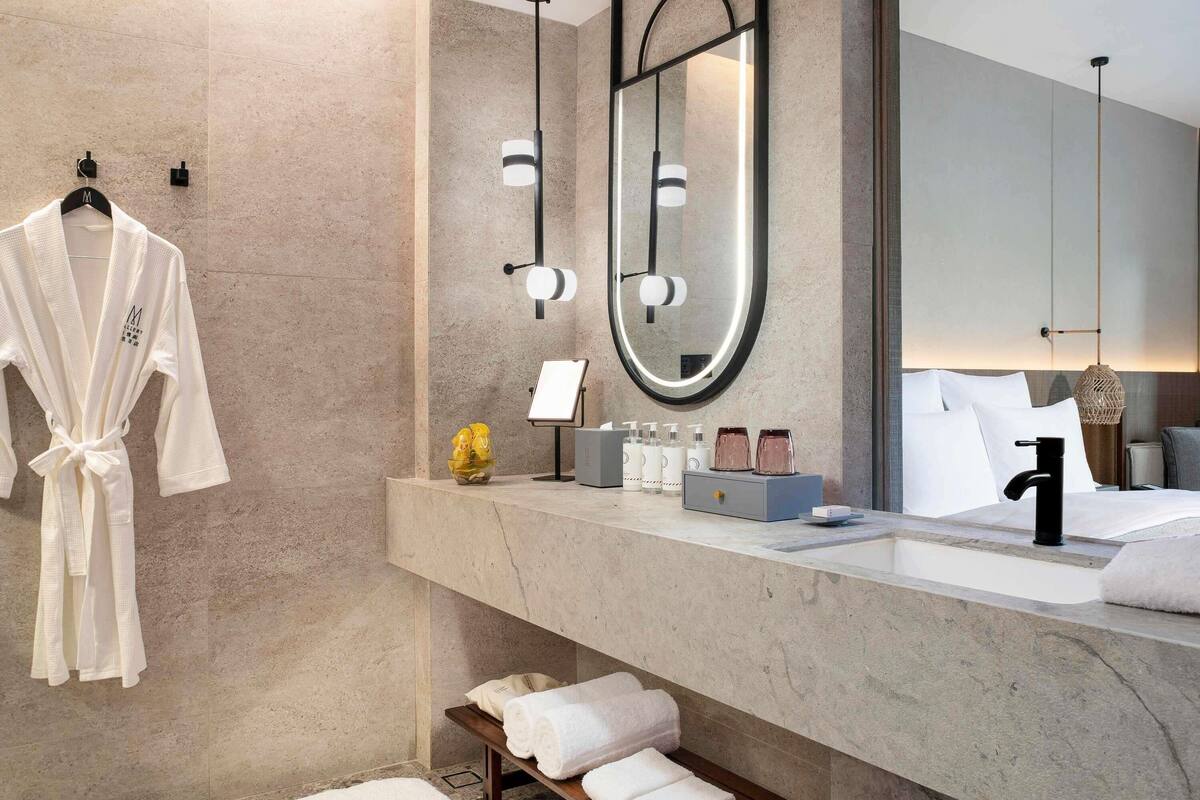 Un baño moderno con encimera de mármol, un espejo grande, una bata colgada en la pared y un banco con toallas.
