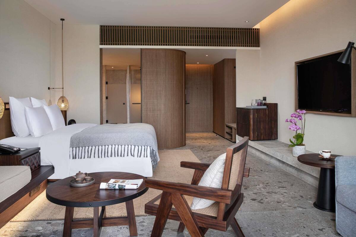A modern hotel room with a bed, bedside table, chair, and a TV mounted on the wall.
