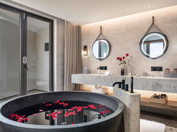 Un baño moderno con una bañera grande, dos espejos, un tocador con lavabo y una amplia cuenca circular adornada con pétalos rojos.