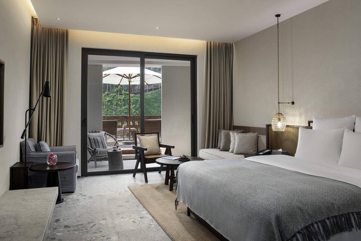 A modern hotel room with a large bed, a desk, a chair, and a view of greenery through a glass door.