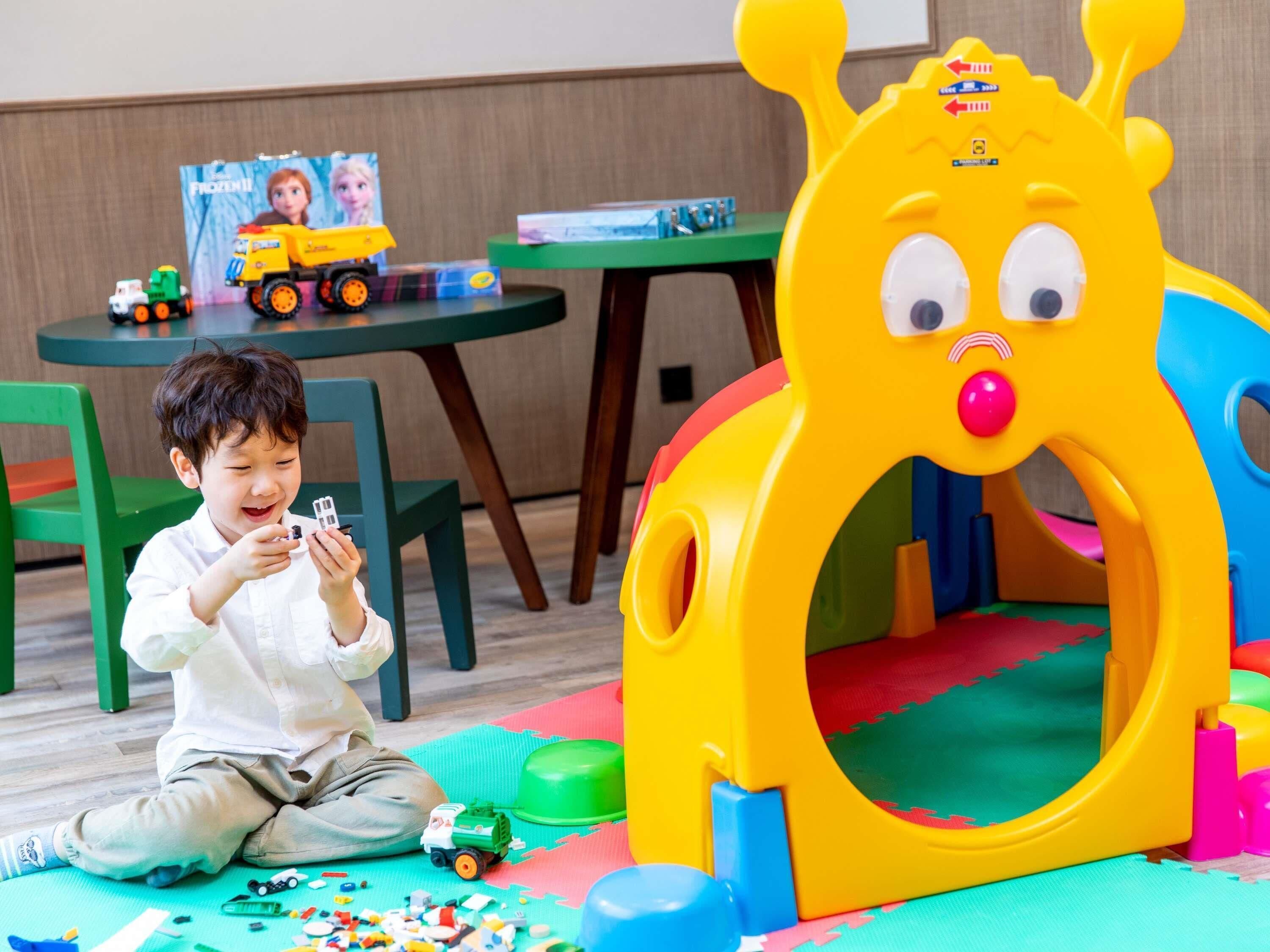 Niño jugando con juguetes en una habitación con una silla verde, una pequeña mesa y una zona de juegos con alfombras y juguetes de colores.