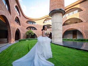 Escena de boda con una novia en vestido blanco y ramo, de pie en un patio con arquitectura de ladrillo y césped verde.