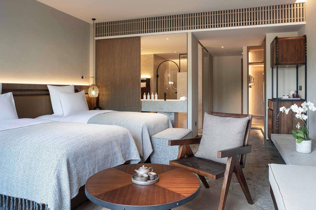 A hotel room with two beds, a round coffee table with a teapot, a chair, and a small table with a potted plant.
