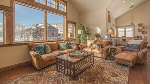 A spacious living room with leather sofas, a coffee table, and large windows offering a view of a cabin.