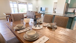 Un coin repas avec une table en bois mise pour quatre personnes, comprenant des bols, des assiettes, des verres et des couverts. On aperçoit un canapé et une cuisine au fond.