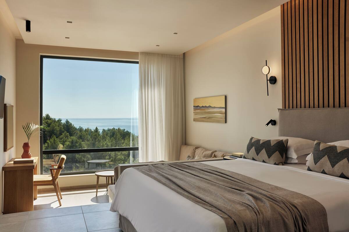 A modern hotel room with a large bed, a view of the ocean, a desk with a chair, and a small table.