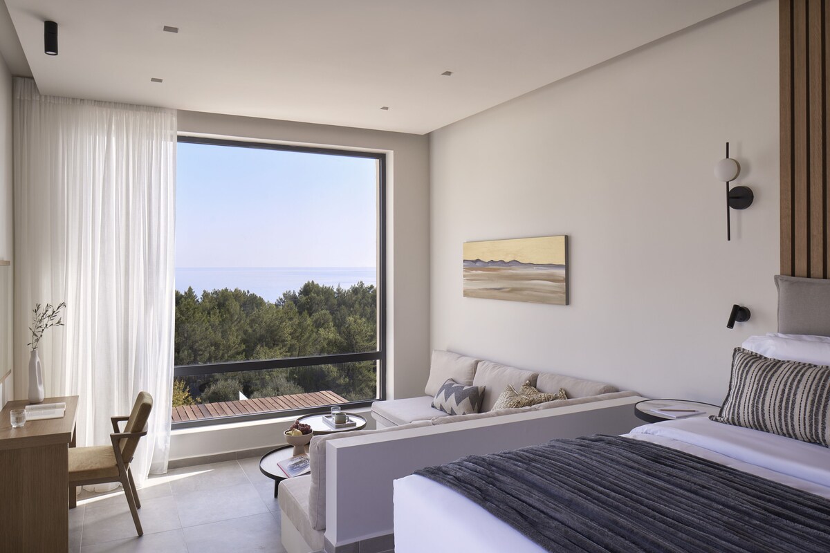 A modern hotel room with a large window, a sofa, a bed, and a desk.