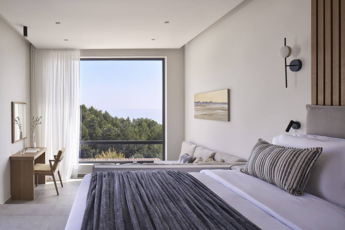 A modern bedroom with a large bed, a desk, and a chair. There is a window with a view of trees and a body of water.