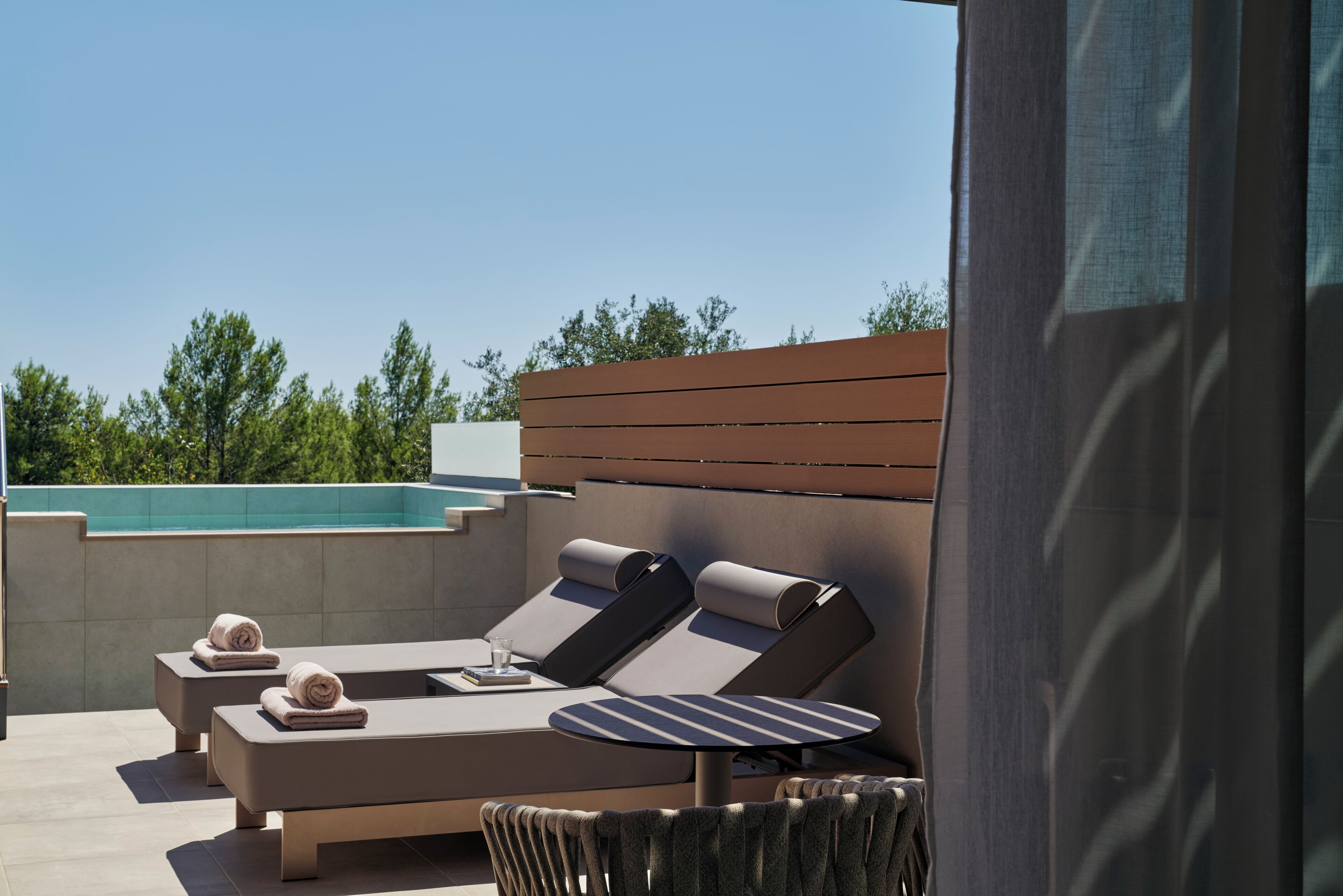 A rooftop lounge area with sun loungers, a small table, and a view of a swimming pool.