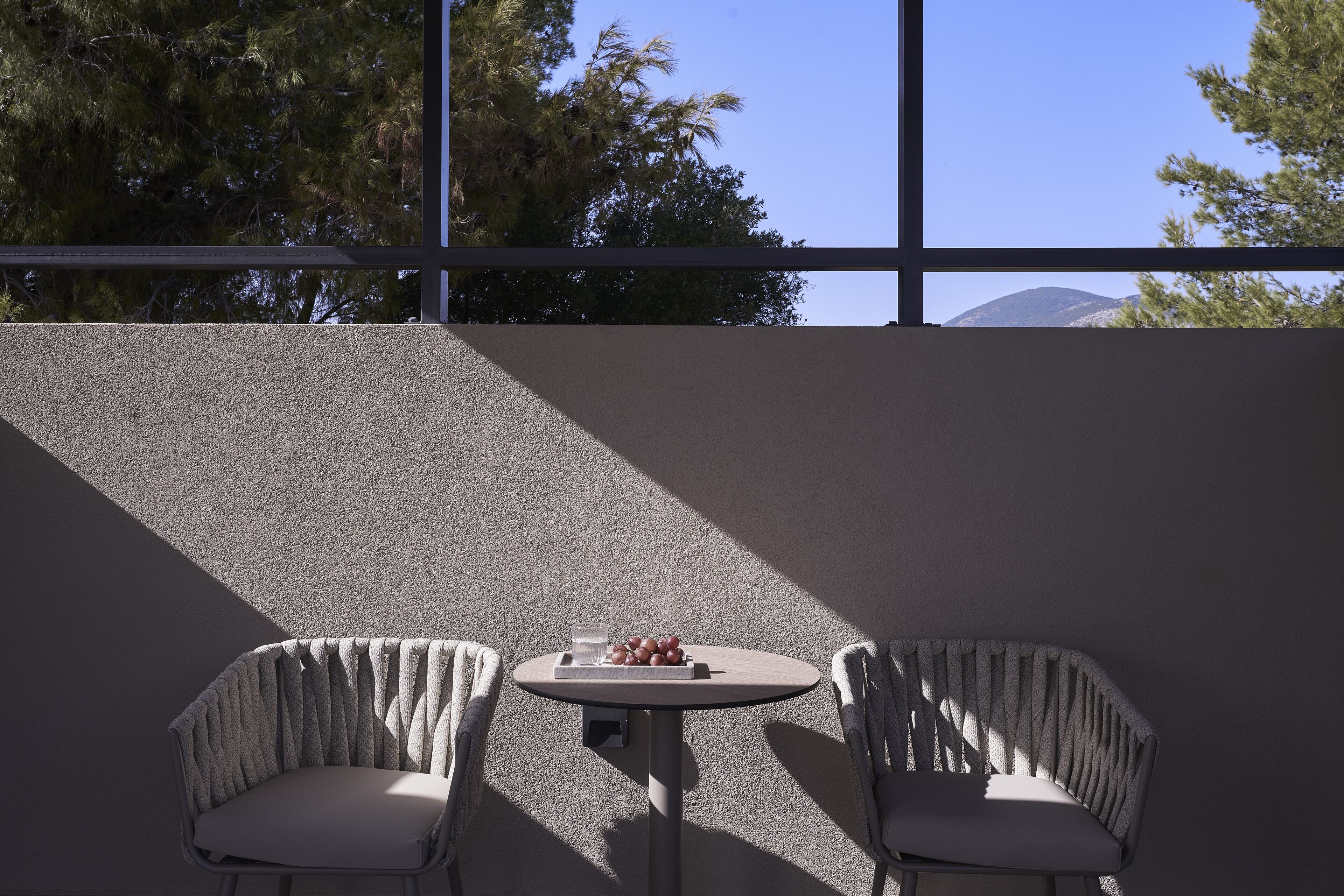 Eine Terrasse mit zwei Stühlen und einem Tisch, Blick auf Bäume und einen Berg durch große Fenster.