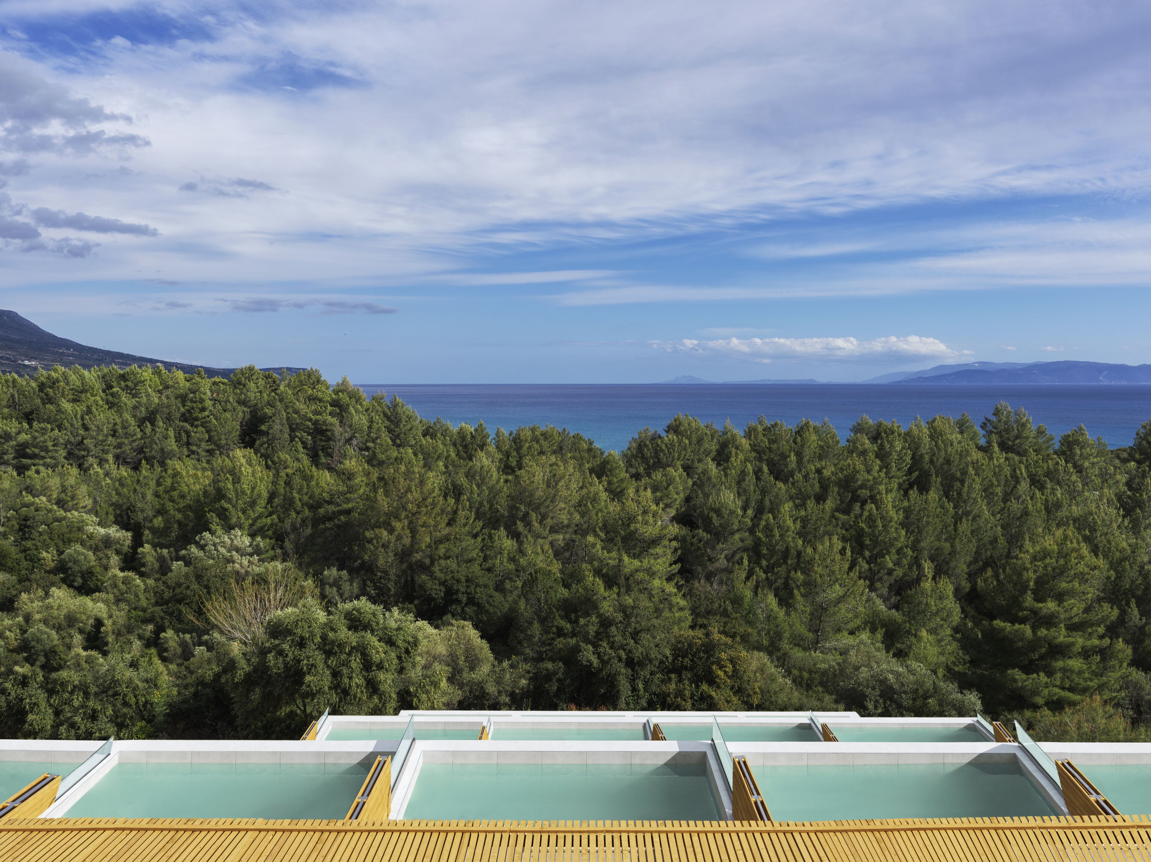 A pool area with multiple pools and a wooden deck, surrounded by dense green trees and a view of the sea and distant mountains.