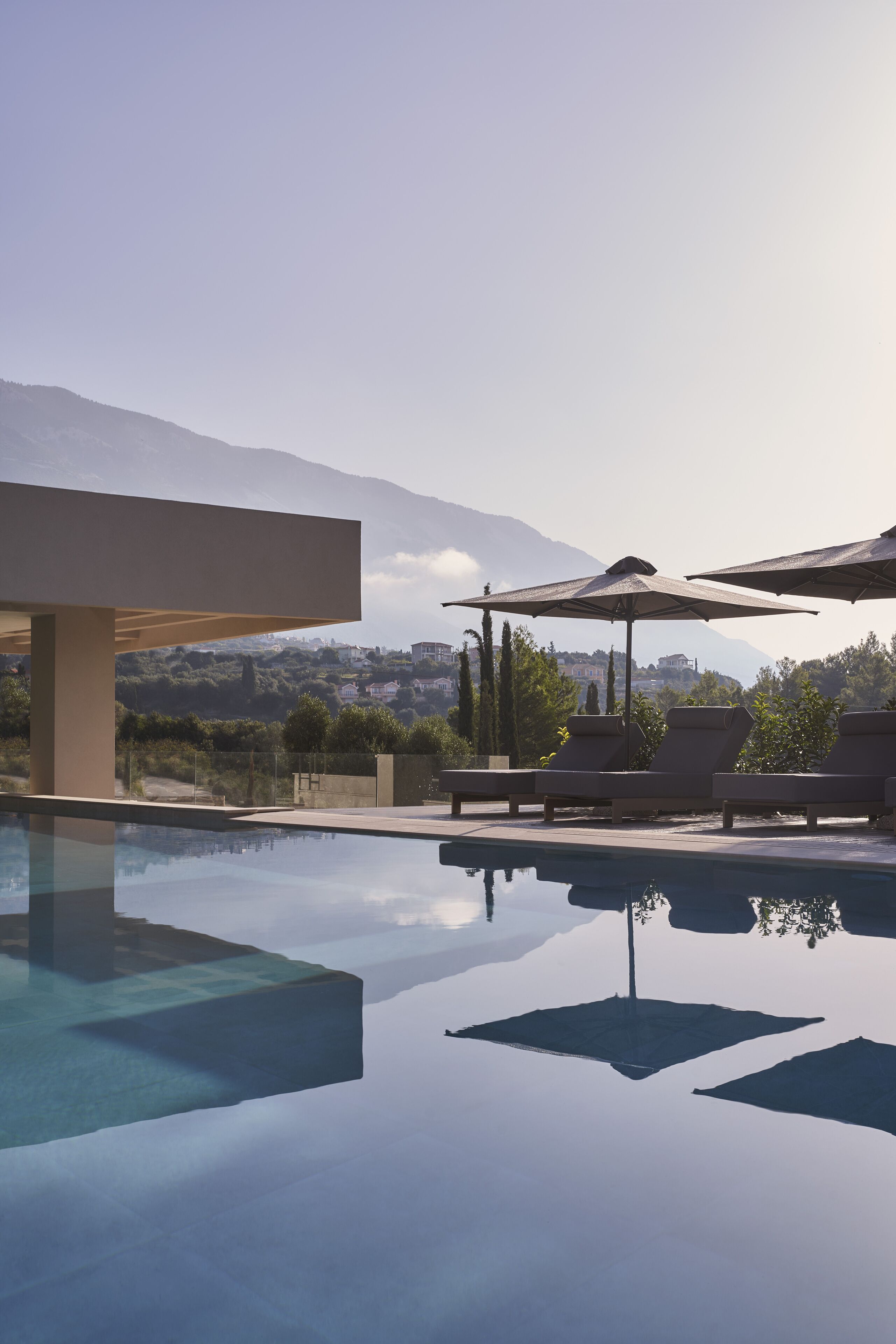 A clear pool with lounge chairs and umbrellas, surrounded by a modern building and a mountainous landscape.