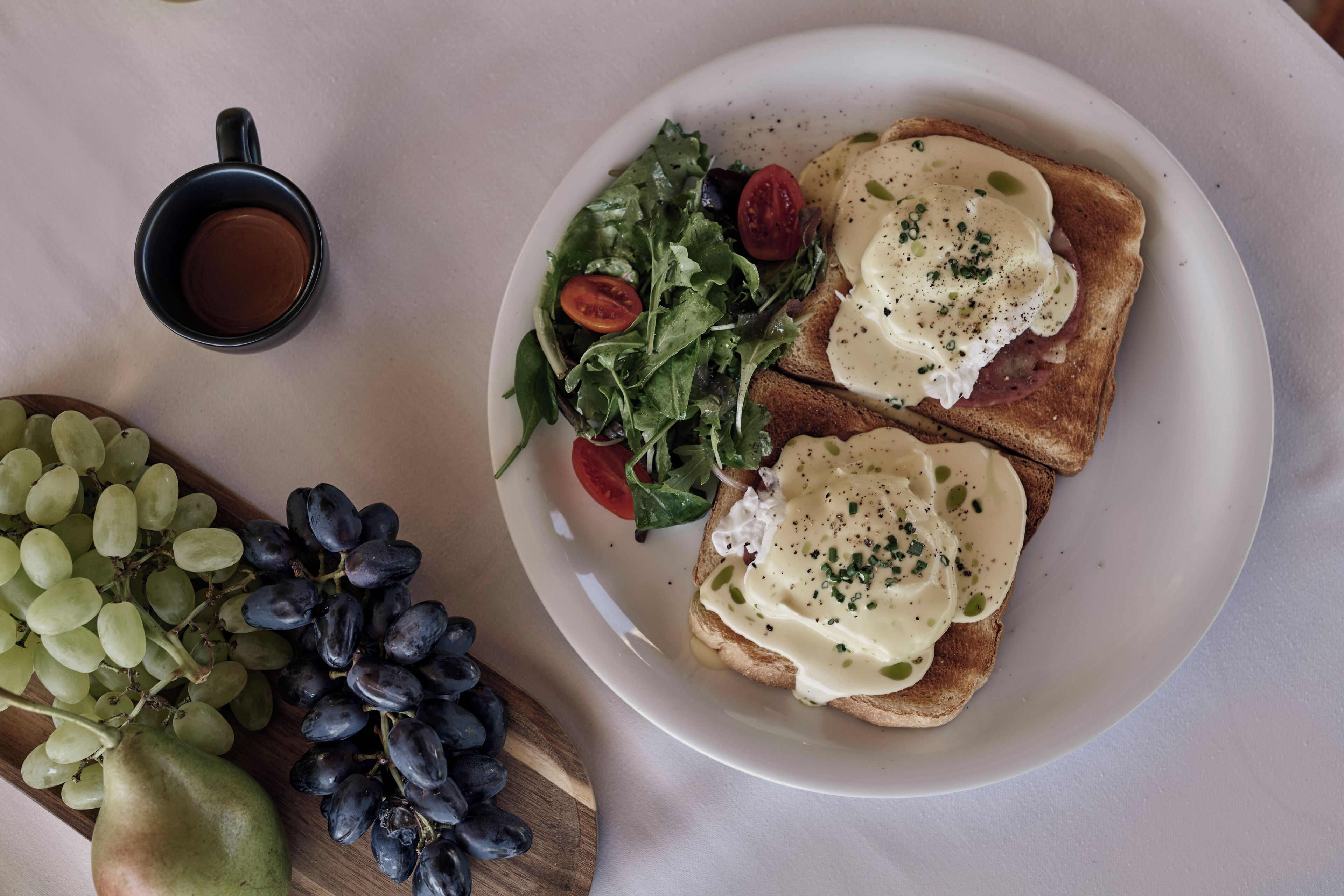 Ein Teller mit Toast, belegt mit Ei und Salat, eine Schüssel mit Trauben und eine Tasse Kaffee auf einem Tisch.