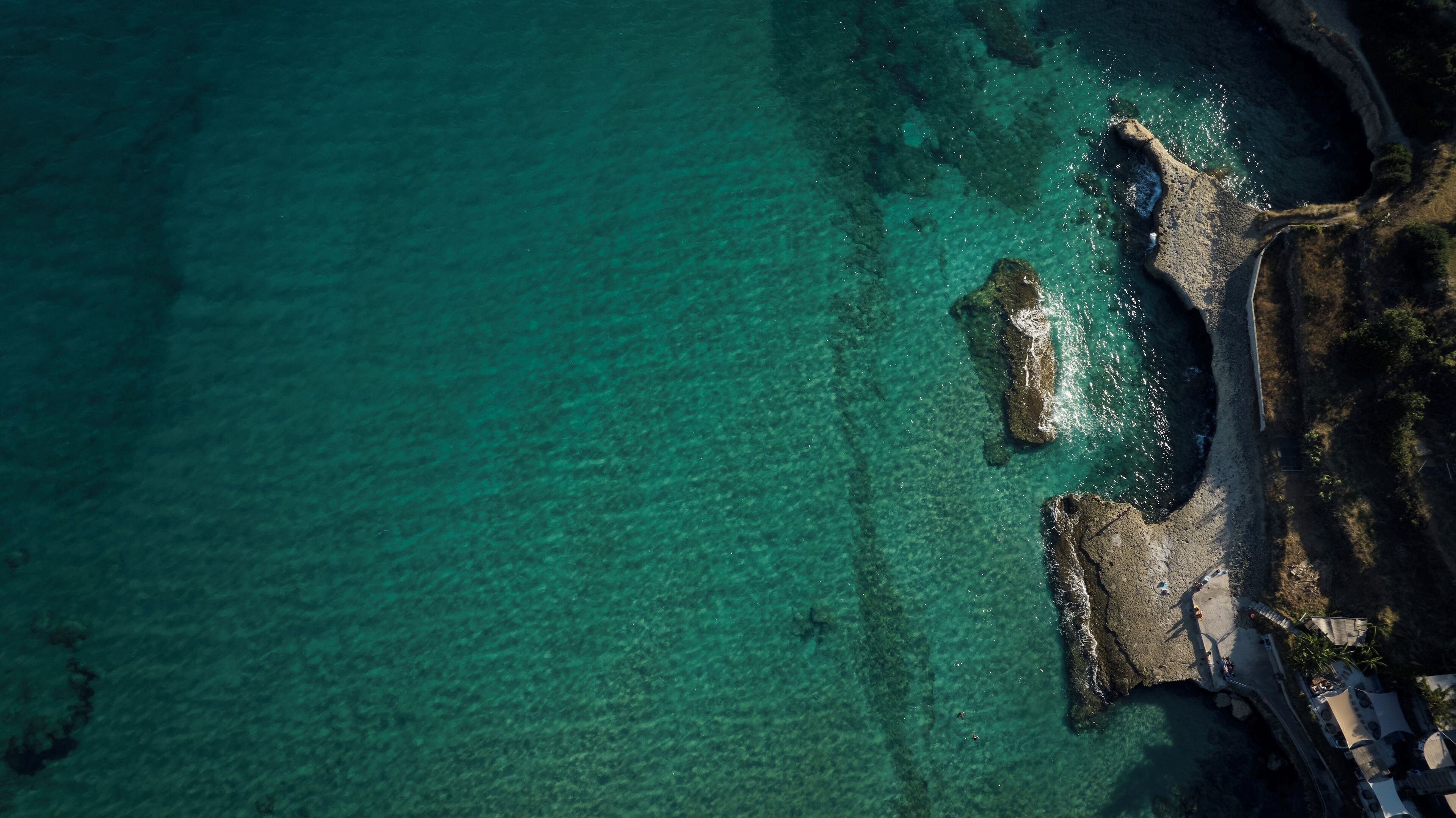 An aerial view of a coastal area with clear turquoise waters, rocky outcrops, and a narrow beach path.