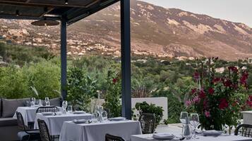 An outdoor dining area with tables set for two, surrounded by greenery and overlooking a mountainous landscape.