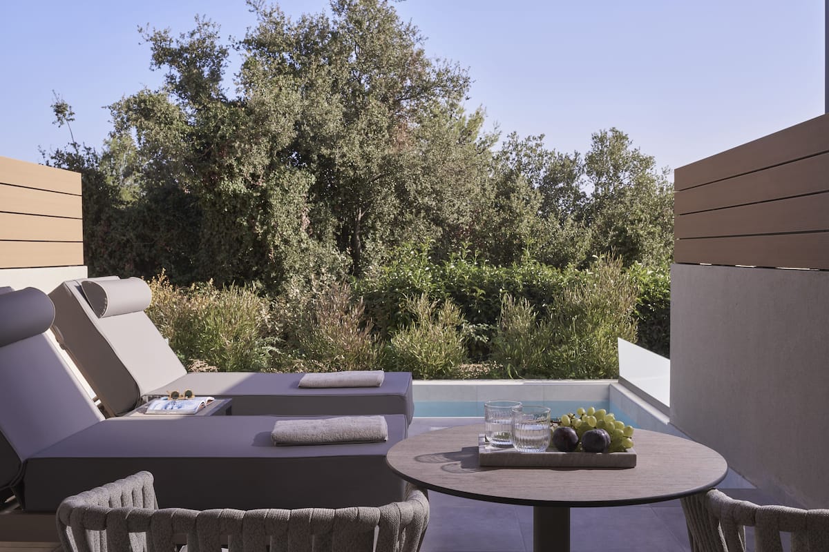 A sunlit outdoor lounge area with a pool, cushioned seating, and a table with a fruit bowl and glasses.