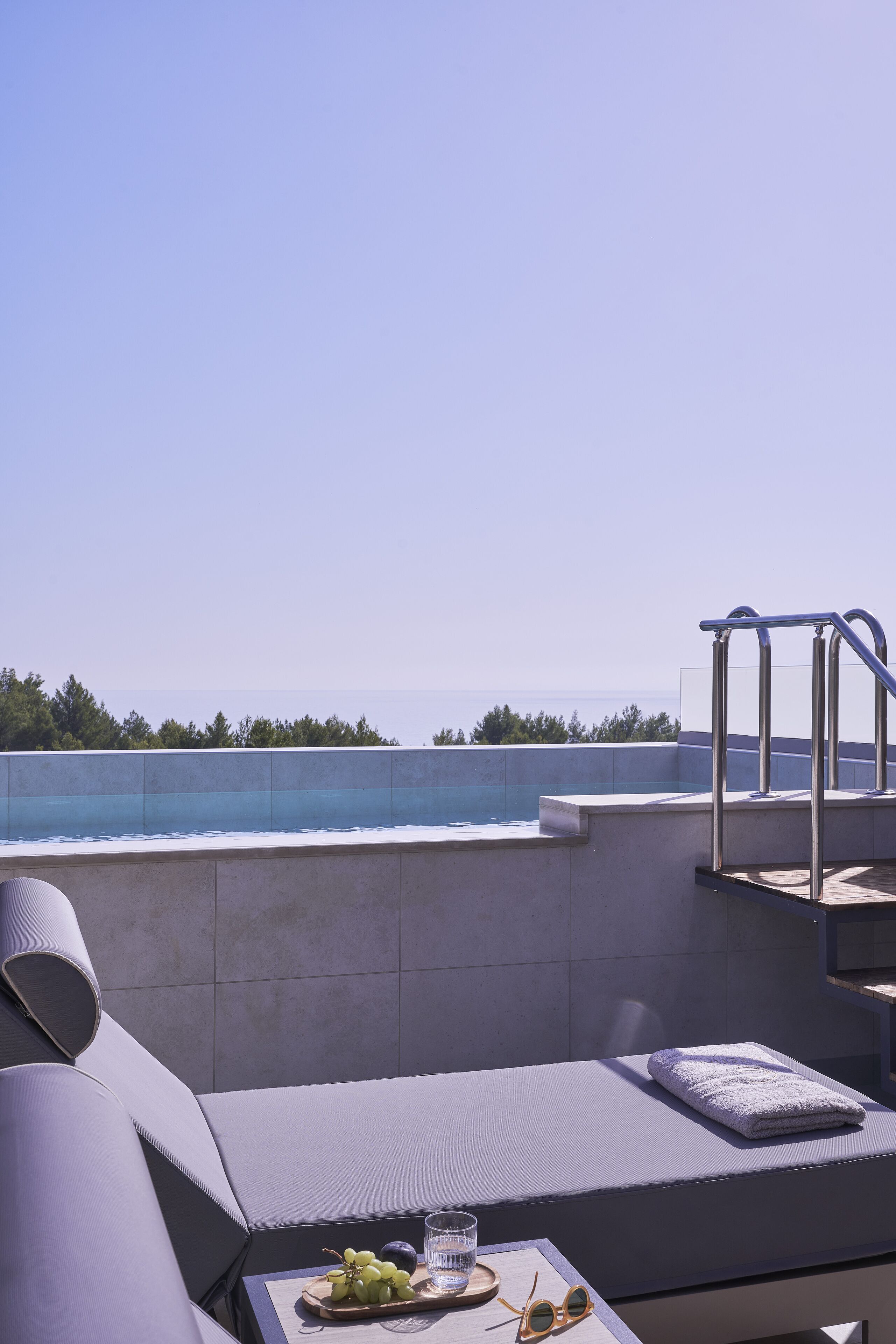 A sunlit balcony with a view of the sea, a cushioned seating area, and a table with a glass of water and grapes.
