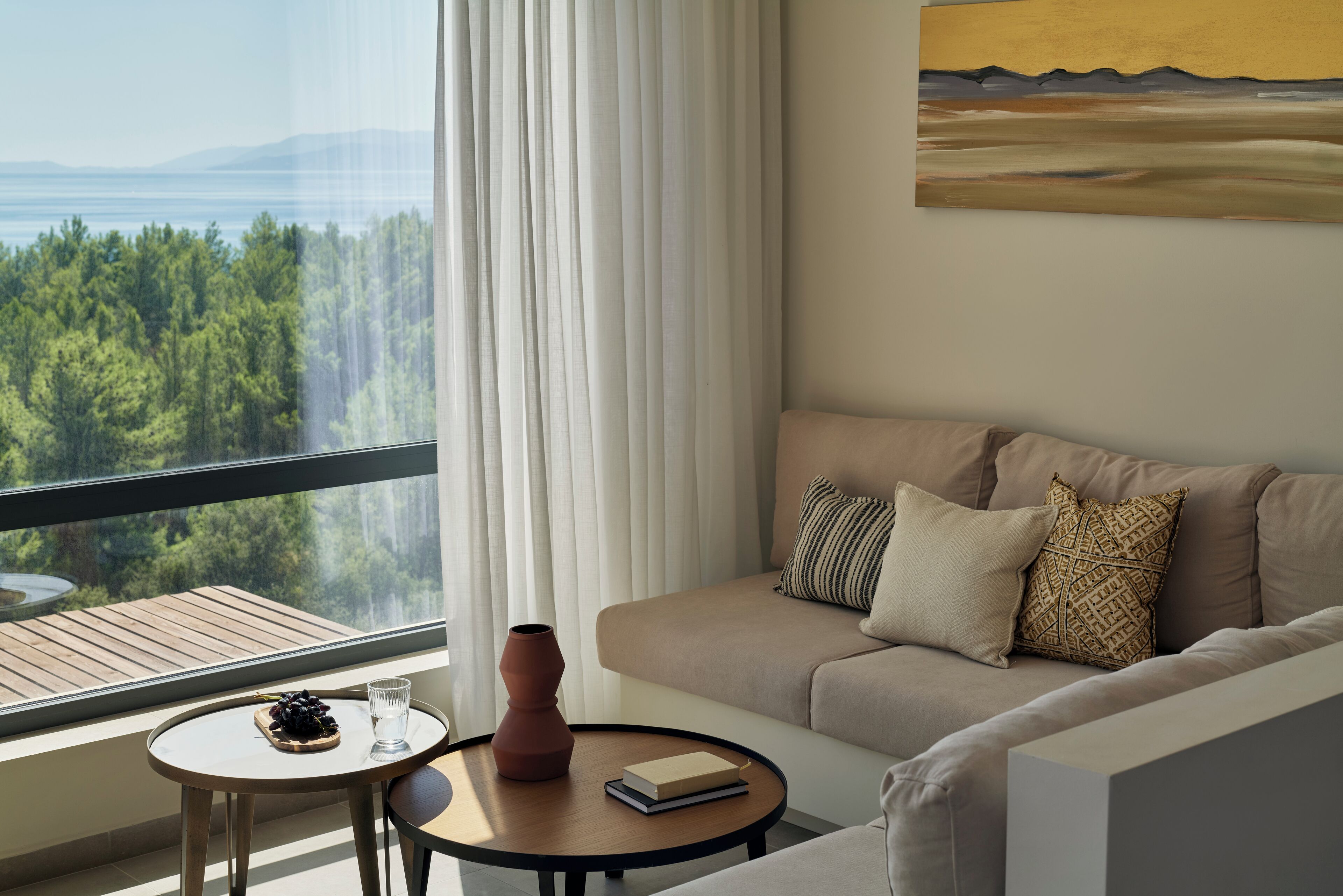 A modern living room with a sofa, a round coffee table, and a view of a forest through a large window.