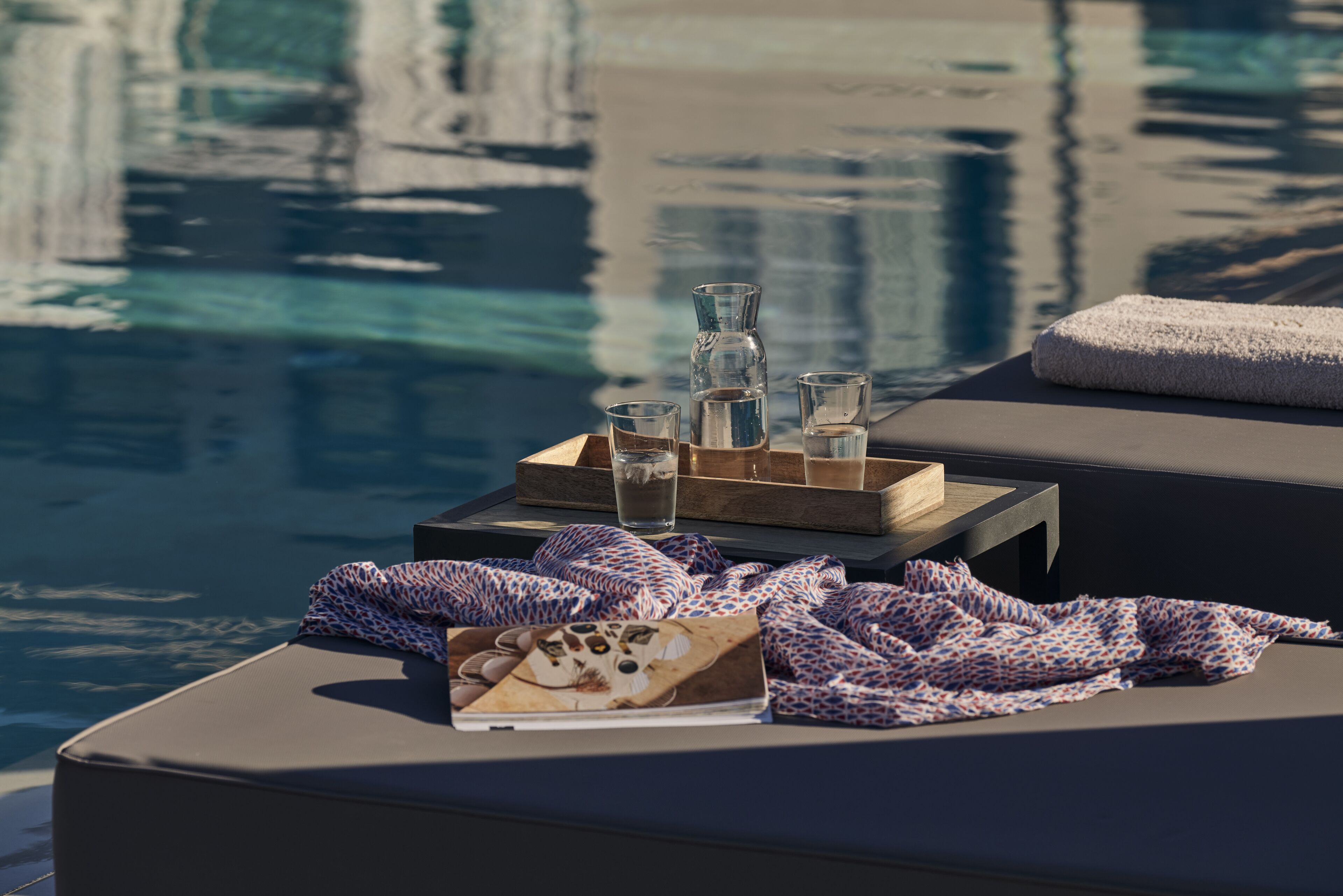 Escena junto a la piscina con una bandeja de vasos y una botella, un textil estampado y un libro sobre una superficie acolchada.