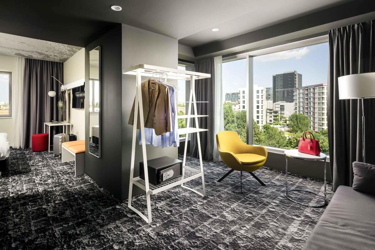 A modern hotel room with a large window, a red ottoman, a yellow armchair, a white coat rack, and a safe.