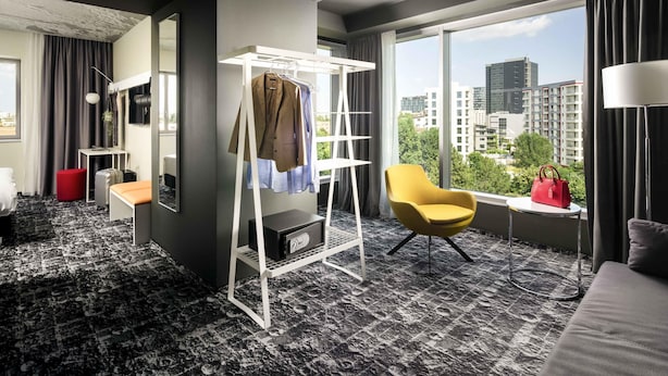A modern hotel room with a large window, a red ottoman, a yellow armchair, a white coat rack, and a safe. - ibis Bucharest Politehnica (Bucharest)