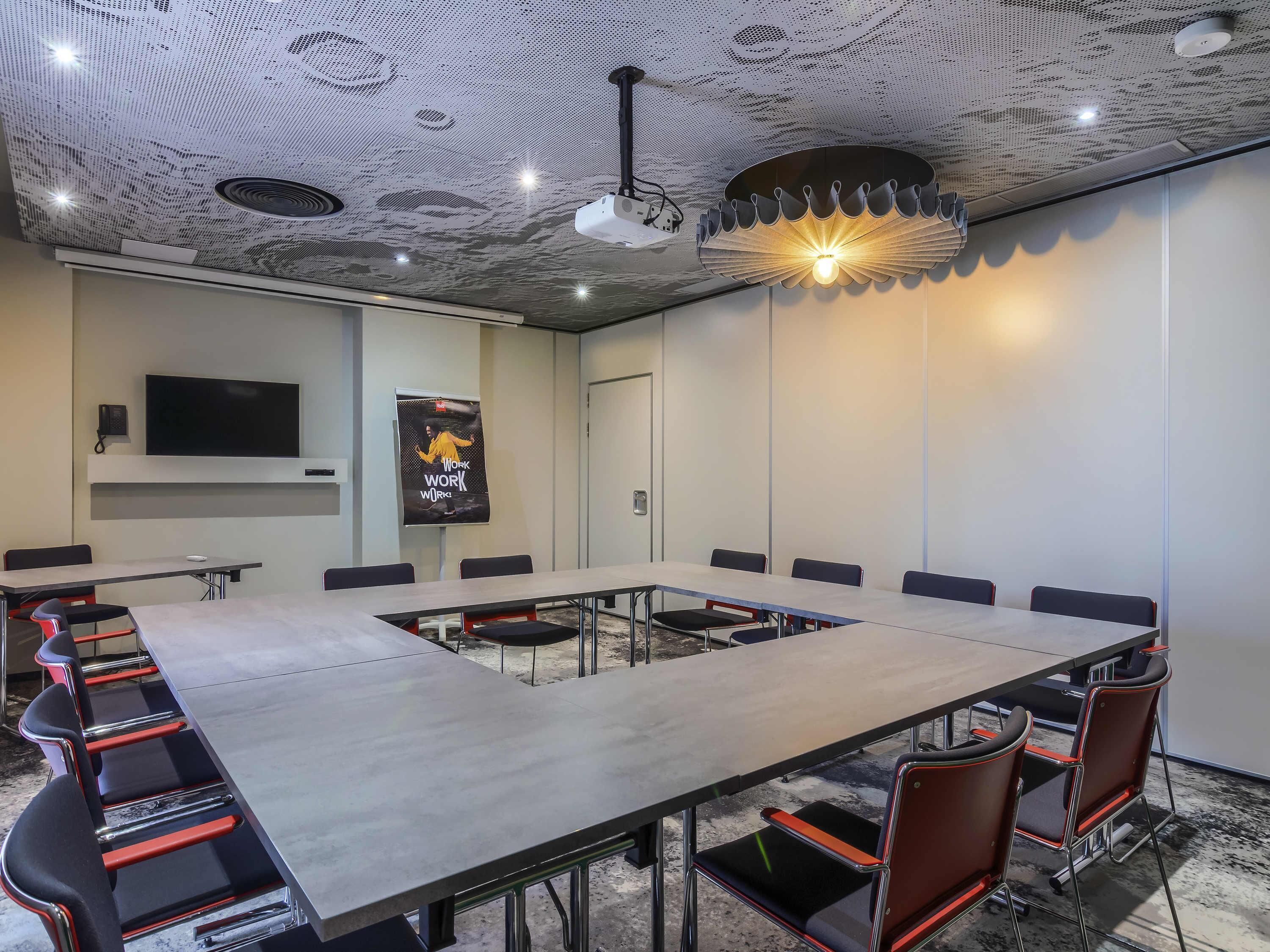 Un salón de conferencias con una larga mesa, sillas, una pantalla de proyector y una lámpara decorativa en el techo.