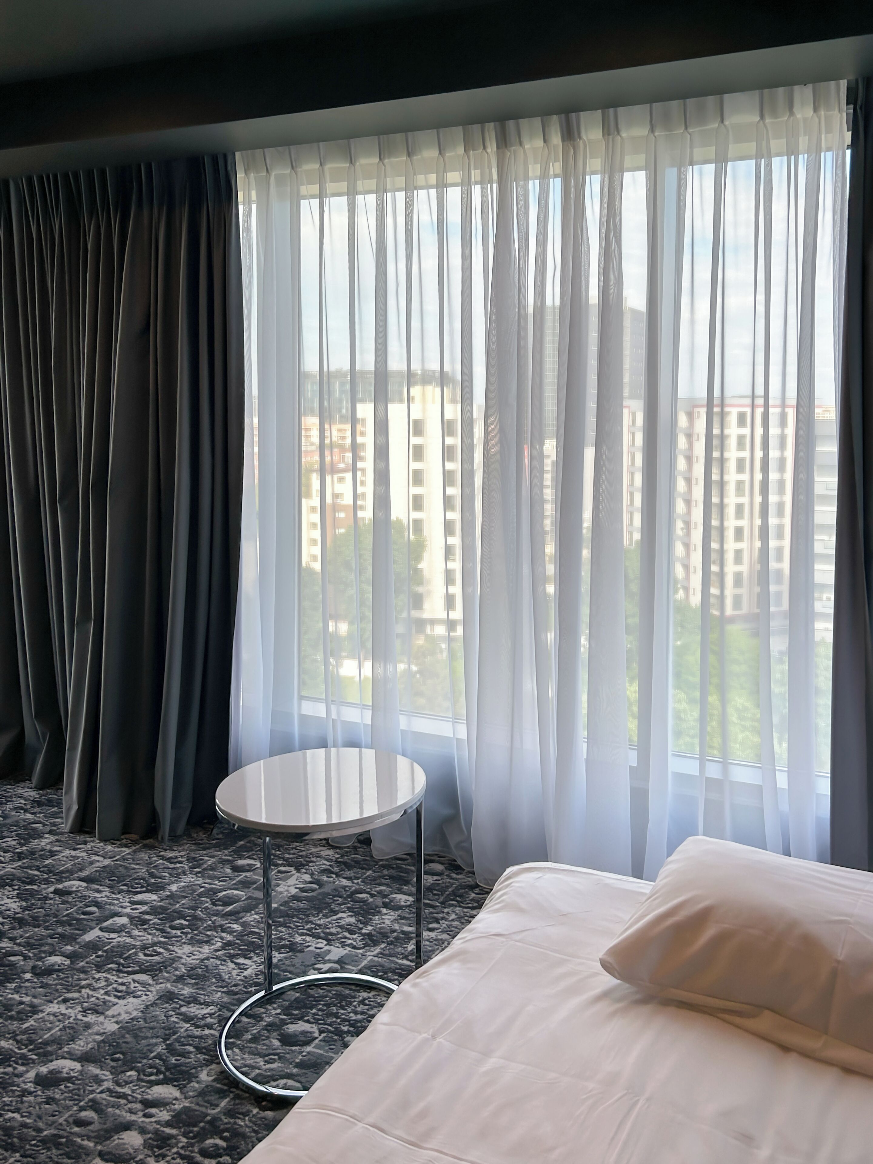Habitación de hotel con cama, una mesita redonda y vistas a edificios a través de cortinas translúcidas.