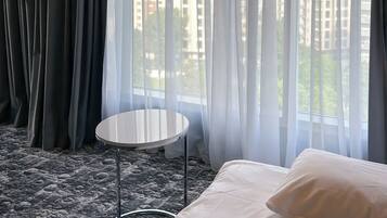 Habitación de hotel con cama, una mesita redonda y vistas a edificios a través de cortinas translúcidas.