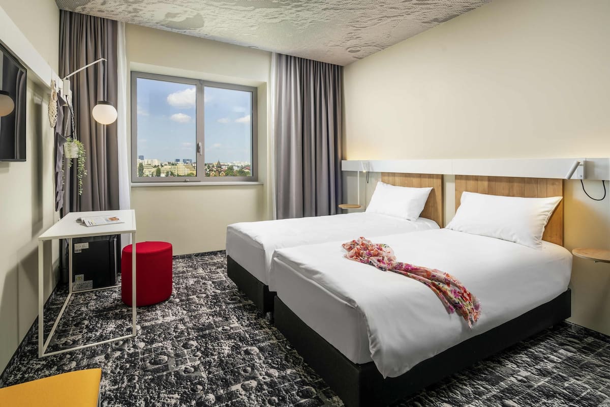 A hotel room with a large bed, a small desk, a red stool, a window with a city view, and a patterned carpet.