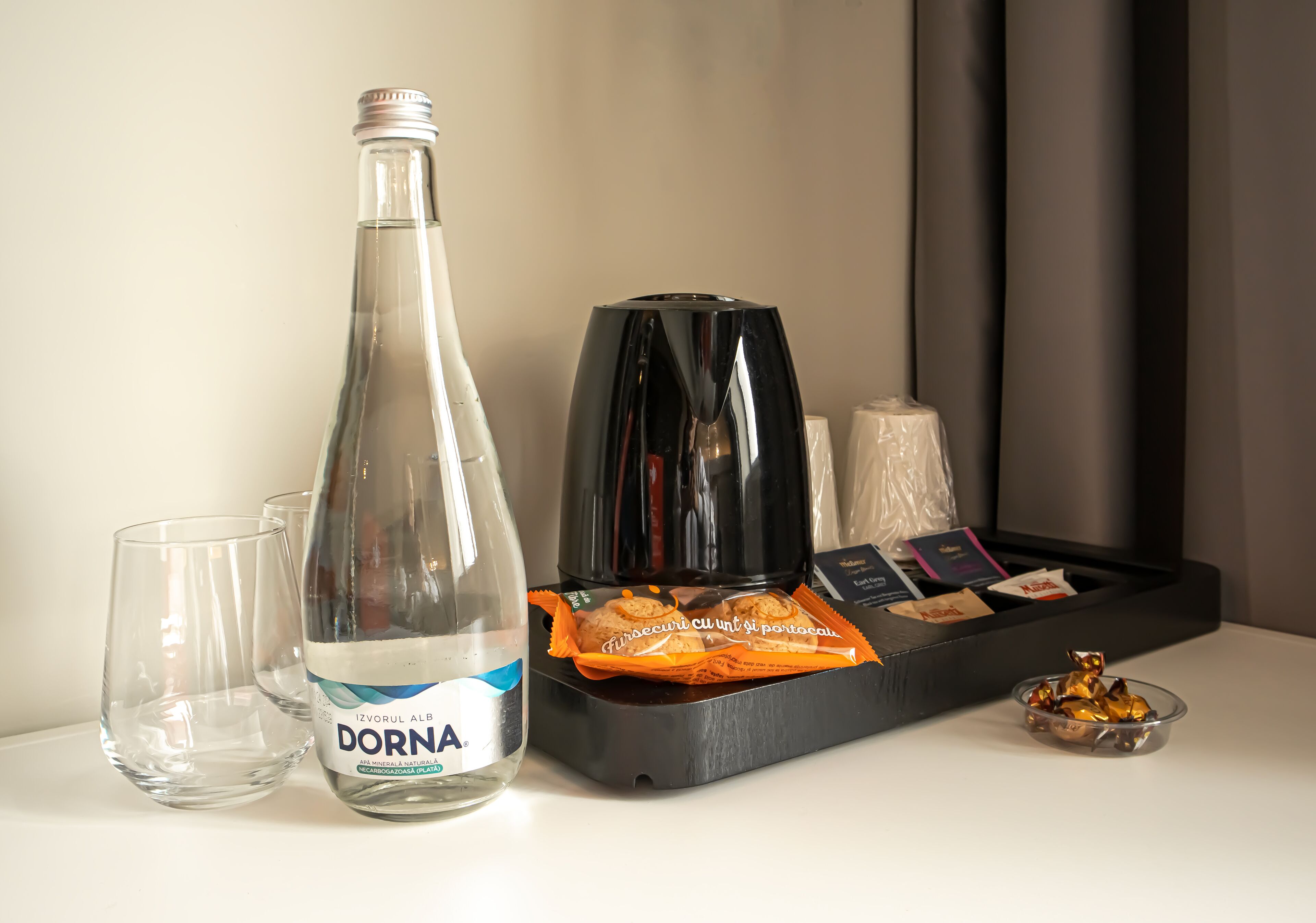 Eine Flasche Dorna Wasser, zwei Gläser, eine Kaffeemaschine, eine Serviertablett mit Snacks und eine kleine Schale Pralinen auf einem Tisch.