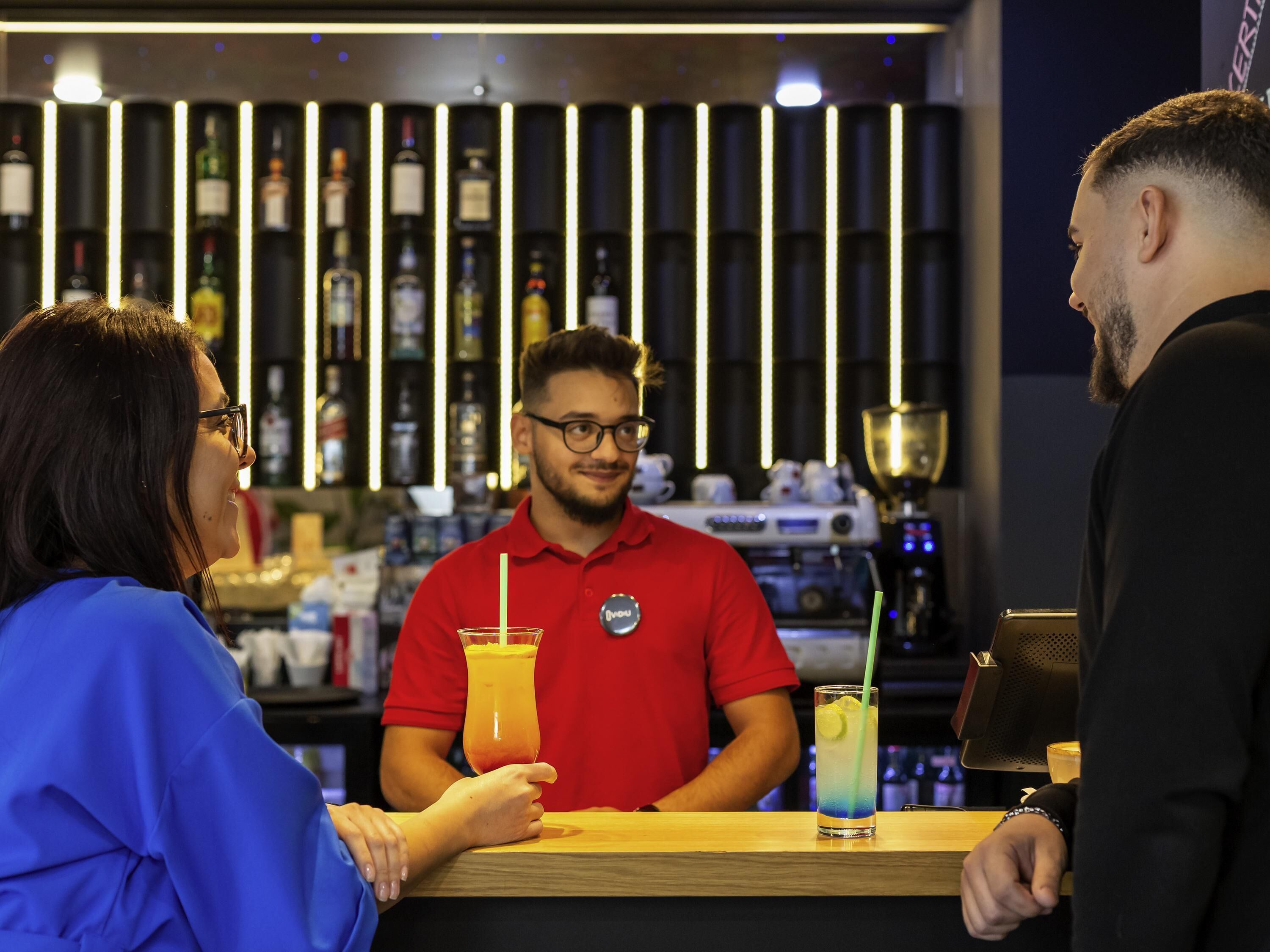Un bar con un camarero con camisa roja, un cliente con camisa azul y otro cliente con camisa negra. Se ven botellas y una máquina de café al fondo.