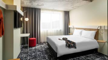 A hotel room with a bed, a TV, a red ottoman, a window with curtains, and a patterned carpet.