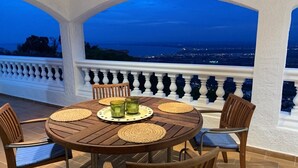 A round wooden table with woven placemats, two glass candle holders, and four wooden chairs with cushioned seats. The table is set against a white balustrade overlooking a cityscape at dusk. - Holiday house in a unique quiet location - sea view - private pool - Internet (Roses , Mas Fumats)