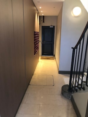Un couloir étroit, avec une porte sombre au fond, un sol carrelé et un escalier doté d’une rampe noire sur le côté droit.