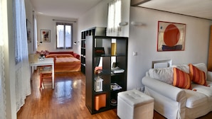 Una sala de estar acogedora con un sofá blanco, una estantería negra, suelo de madera y una ventana con cortinas.