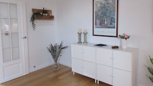 Een kamer met een witte deur, een witte console en een lijst met een schilderij, een vaas met gedroogde planten, en een houten plank met wijnflessen.