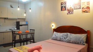 A bedroom with a wooden headboard, a bed with a pink bedspread, and a nightstand with a rolled towel. There is a small table with two chairs and a toaster on the counter. The room has a kitchenette with cabinets and a microwave. - Dencio's Crib (Lapu-Lapu City)