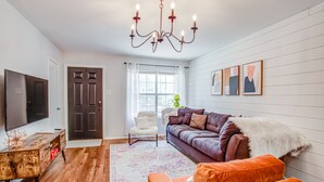 A living room with a wooden floor, a brown sofa, an orange armchair, a white armchair, a TV, and a chandelier. - Fort Worth Hideaway Near Downtown (Fort Worth)