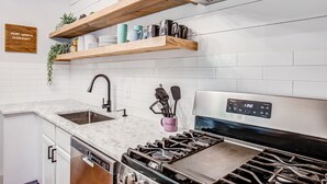 A modern kitchen with white subway tiles, a stainless steel stove, wooden shelves, and a dish rack. - Fort Worth Hideaway Near Downtown (Fort Worth)