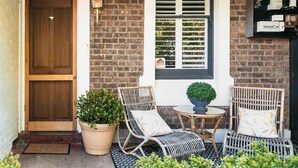 Une façade en briques avec une porte en bois, une fenêtre avec des volets, des sièges à l’extérieur et un pot de fleurs.