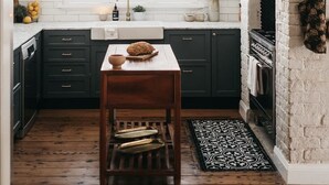 Une cuisine avec des meubles de couleur sombre, une table en bois, un luminaire suspendu et un mur en briques.