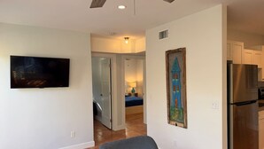 A living room with a ceiling fan, a flat-screen TV, a painting, and a kitchen area with a refrigerator. - Central, Quiet and Beautifully Decorated #1 (Fort Lauderdale)