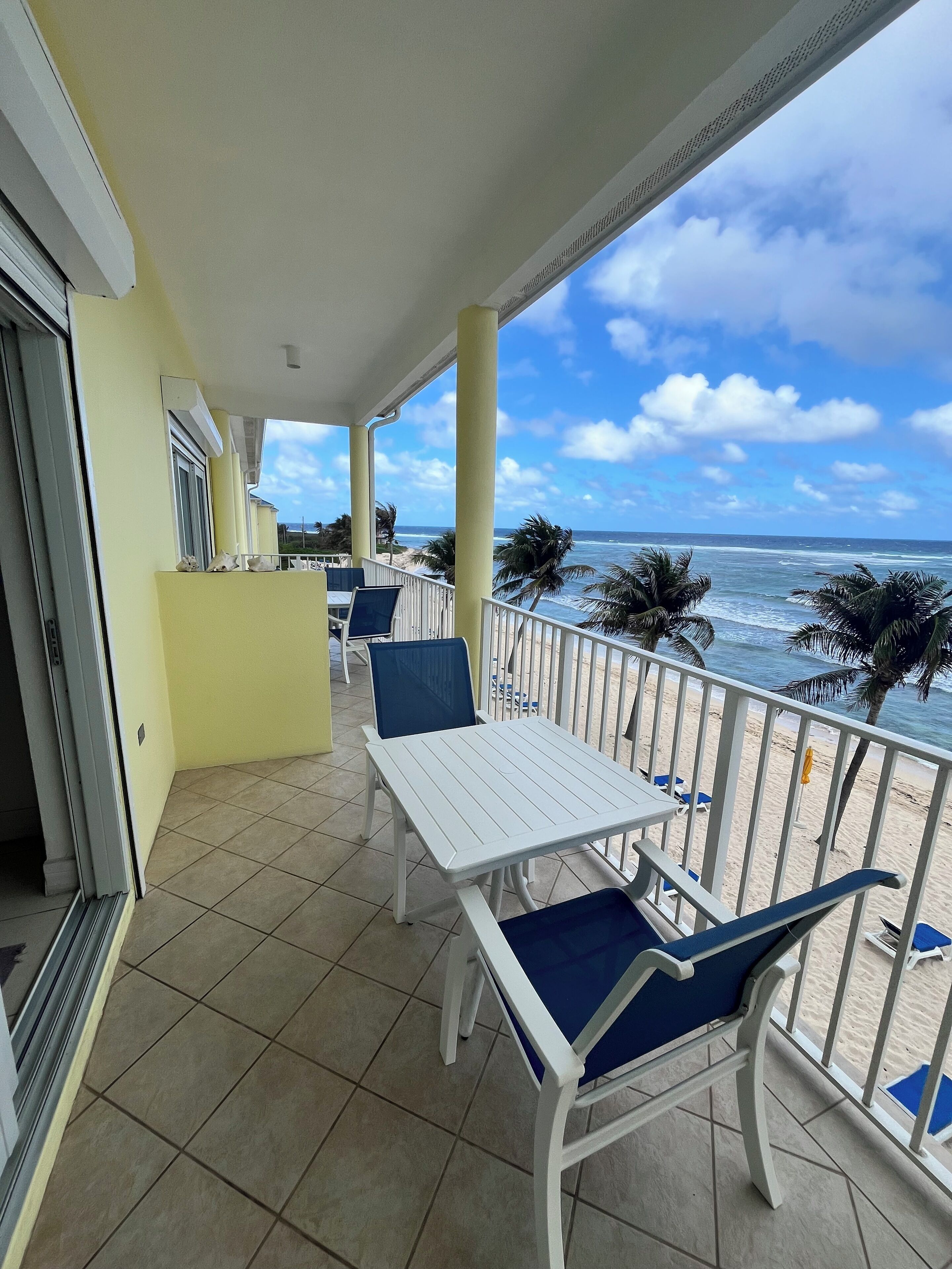 Un balcon avec une table et des chaises blanches, une vue sur la plage et des palmiers.