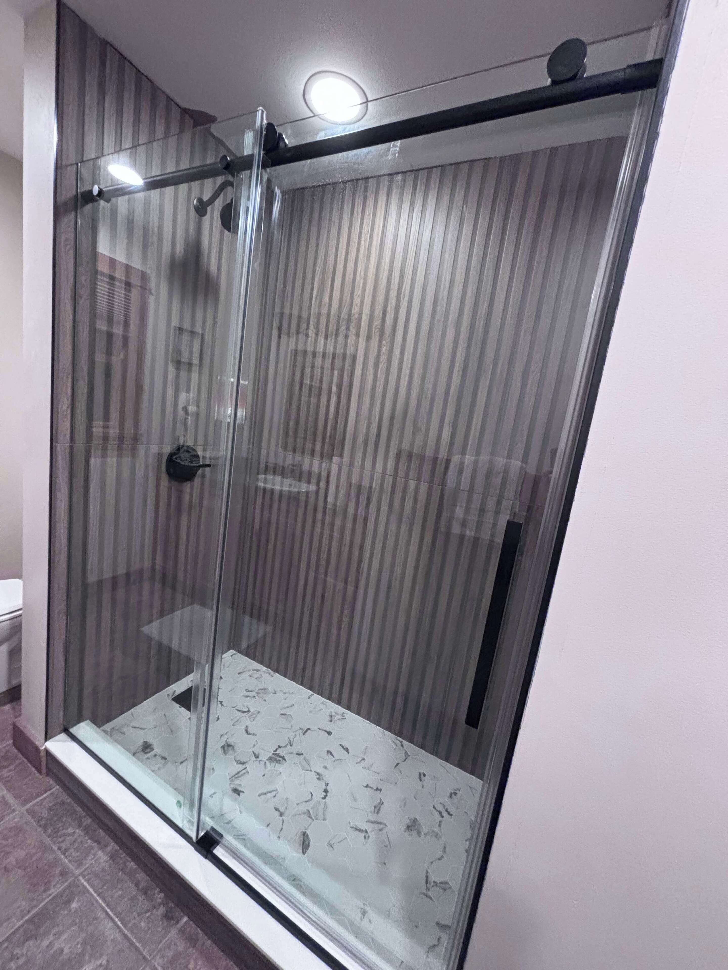 Une douche fermée par des parois de verre, avec un sol carrelé et un mur à rayures verticales.