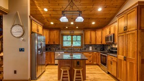 A spacious kitchen with wooden cabinets, a central island with stools, a stainless steel refrigerator, and a large window providing natural light.