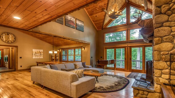 A spacious living room with a wooden ceiling, large windows, a stone fireplace, and a cozy sofa.